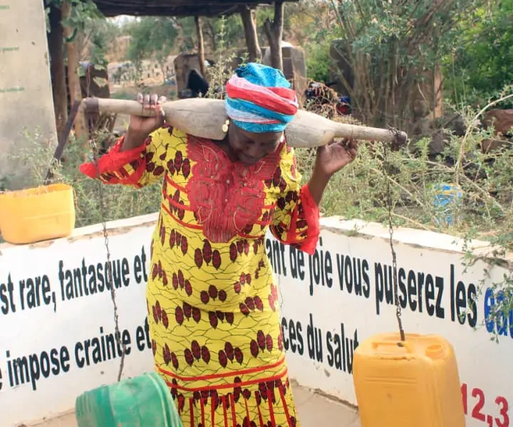 Une femme porte deux bidons à la palanche devant les pancartes du musée.